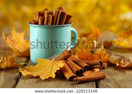 Cinnamon sticks in mug with yellow leaves on table on bright background