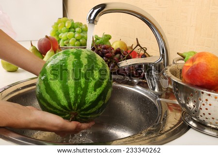 Woman's hands washing watermelon and other fruits in colander in sink