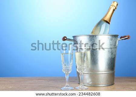 Bottle of champagne in metal ice bucket and two glasses on wooden table on color background