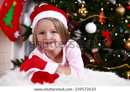Little girl in Santa hat and mittens lying on fur carpet on Christmas tree background