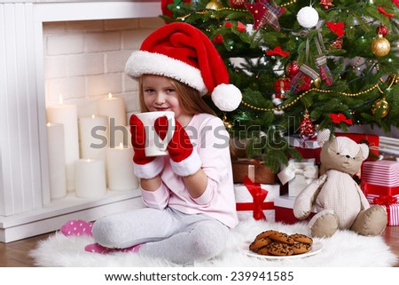 Little girl in Santa hat and mittens taking cup sitting near fir tree on fur carpet and wooden floor, on fireplace with candles background