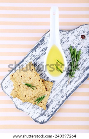 Crispbread with spoon of oil and sprigs of rosemary on cutting board, on striped background