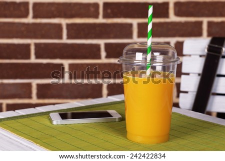 Orange juice in fast food closed cup with tube and mobile phone on wooden table and brick wall background