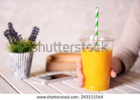 Female hand at wooden table with fast food closed cup of orange juice and near books, plant and mobile phone on light wall background