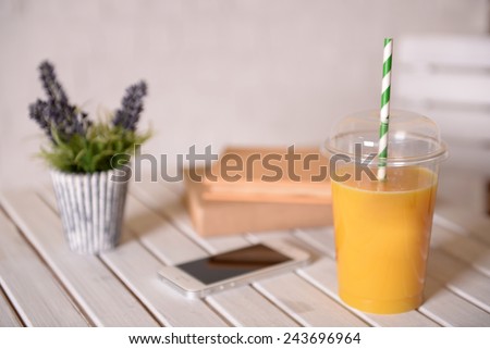 Orange juice in fast food closed cup with tube on wooden table with plant and light wall background