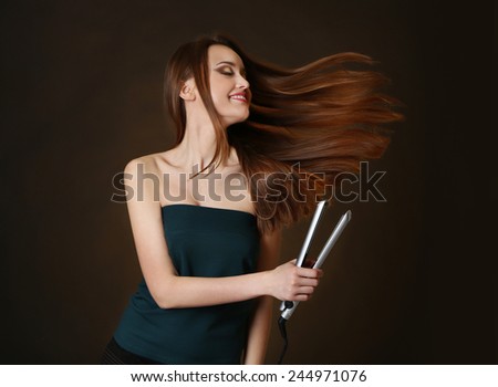 Beautiful young woman with long hair using hair straighteners on dark brown background