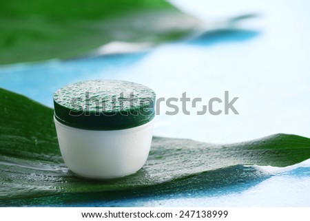 Cosmetic cream with water drops on green leaves on light background