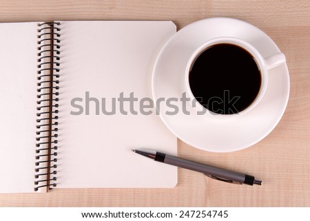Cup of coffee on saucer with notebook and pen on wooden table background