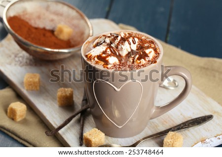 Cup of hot coffee with marshmallow on cutting board with sieve of cocoa, lump sugar and sticks of vanilla on color wooden table background