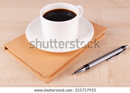 Cup of coffee on saucer with close notebook and pen on wooden table background