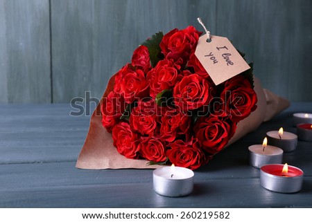 Bouquet of red roses with tag wrapped in paper and candles on wooden background