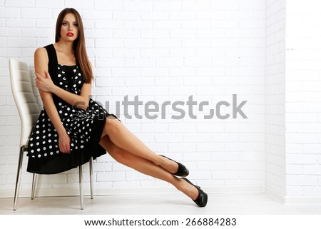 Young woman sitting on chair in dots dress on white brick wall background