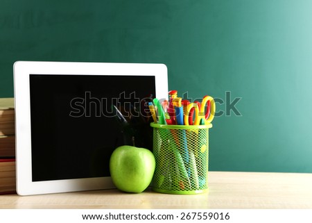 Digital tablet, books, colorful pens and apple on desk in front of blackboard