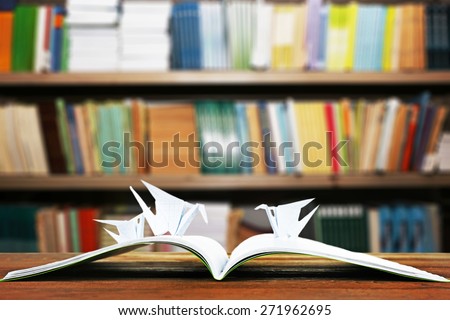 Open book with paper cranes on bookshelves background