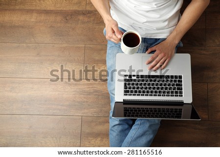 Young man sitting on floor with laptop and cup of coffee in room