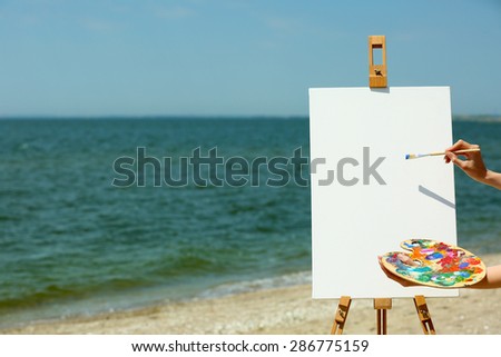 Female hand holding palette with paints and easel with canvas on beach