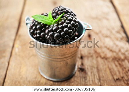 Ripe blackberries with green leaves in small metal bucket on wooden background