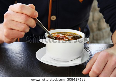 White bowl with borsch and male hands with spoon on wooden table, closeup