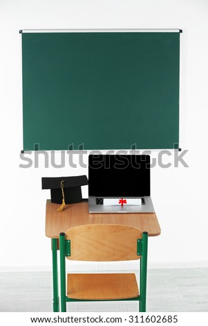 School desk with laptop, master hat and diploma in classroom
