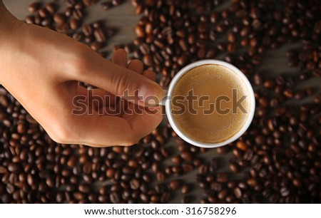 Female hand holding cup of coffee with foam over grains, top view