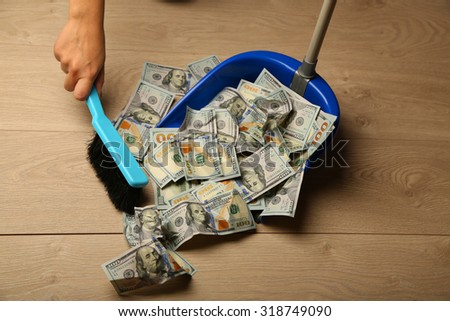 Woman broom sweeps dollars in garbage scoop on wooden floor background