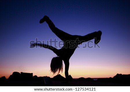 Silhouette of an athletic teenager breakdancing at sunset silhouetted against the colourful sky balance upside down on one hand while cartwheeling