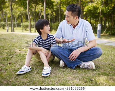 asian father and elementary-age son sitting on grass outdoors having a serious conversation.