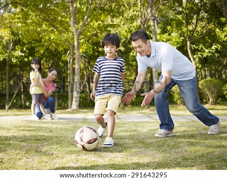 asian father teaching son to play soccer (football) in a park while mother and daughter watching from behind.