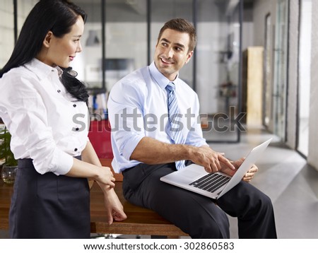 caucasian businessman pointing  to laptop screen while talking to his female asian coworker in office of a multinational company.