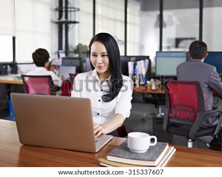 young asian businesswoman working in office using laptop computer with colleagues in background.