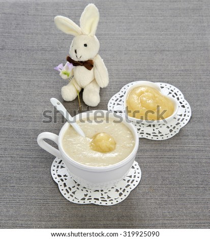 baby nutrition porridge with apple in a white cup on a gray linen table cloth with a toy bunny in the background