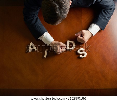 Word AIDS made of wooden block letters and devastated middle aged caucasian man in a black suit sitting at the table, top view composition with dramatic lighting
