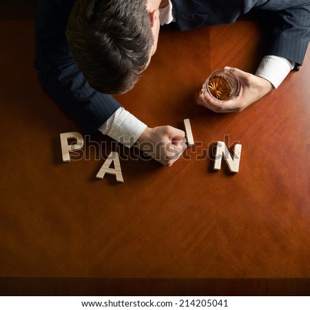 Word Pain made of wooden block letters and devastated middle aged caucasian man in a black suit sitting at the table with the glass of whiskey, top view composition with dramatic lighting