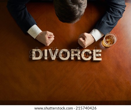 Word Divorce made of wooden block letters and devastated middle aged caucasian man in a black suit sitting at the table with the glass of whiskey, top view composition with dramatic lighting
