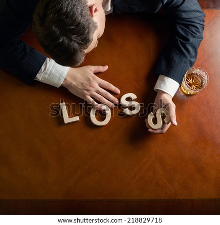 Word Loss made of wooden block letters and devastated middle aged caucasian man in a black suit sitting at the table with the glass of whiskey, top view composition with dramatic lighting