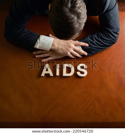 Word AIDS made of wooden block letters and devastated middle aged caucasian man in a black suit sitting at the table, top view composition with dramatic lighting