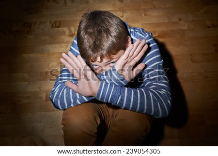 Child abuse composition of a frightened young boy sitting on the wooden floor in a light of a flashlight circle