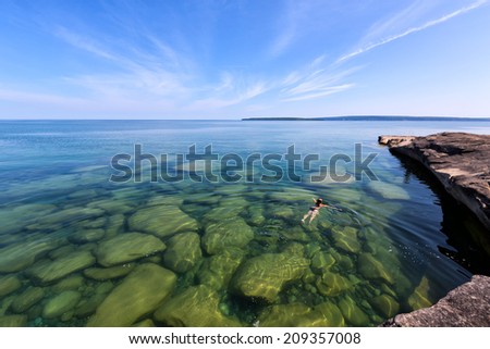 Girl swims in Lake Superior in the Upper Peninsula of Michigan. Rocks are visible through the glass like, pristine waters. Pictured Rocks National Lakeshore is in the distance.