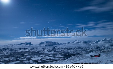 The star sign Orion above a moonlit mountain landscape with a single cozy house lower right.