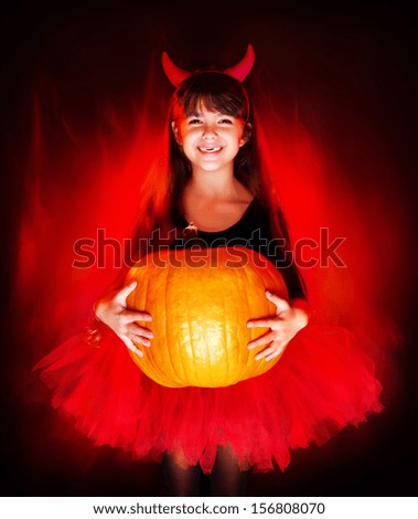 smiling little halloween devil girl holding big pumpkin on fire background