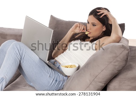 young woman lying on the couch with a laptop in front of her and eating potato chips