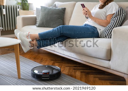 Robot vacuum cleaner cleaning the living room. Young woman enjoy rest, sitting on sofa at home