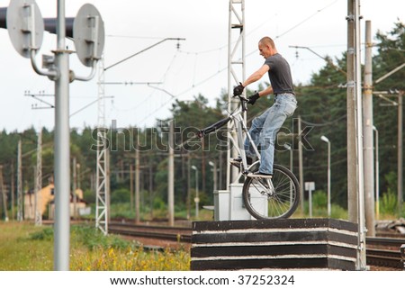 Young man standing on concrete blocks without front wheel in urban area with railways in background. Low angle view.