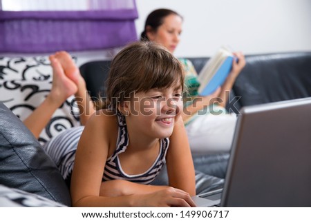 Little girl using computer in the living room while her mother is reading a book