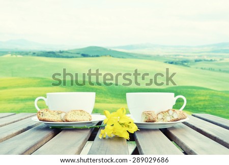 Filtered image of two coffee cups and cantuccini on the wooden table against Tuscan landscape, Italy