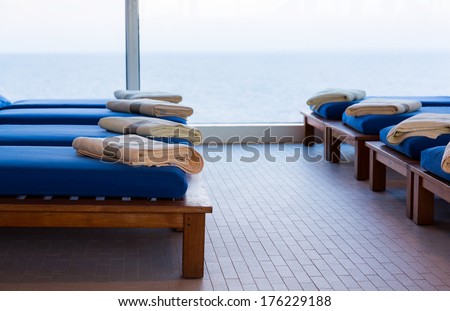 Row of teak reclining couches or benches with cushions and towel by window looking out to sea with horizon in the distance