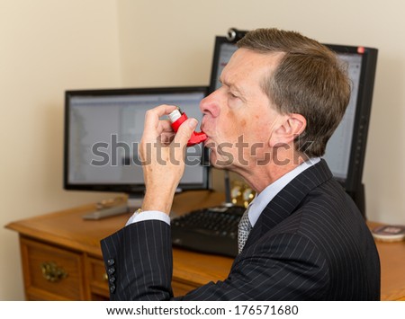 Senior caucasian man  in suit at desk with computer screens with asthma inhaler to handle problems with breathing