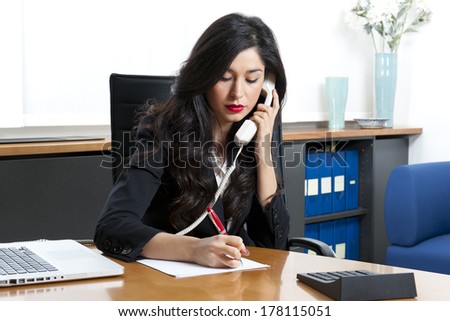 Young business woman talking at the phone writing on the notepad sitting at the desk on office background