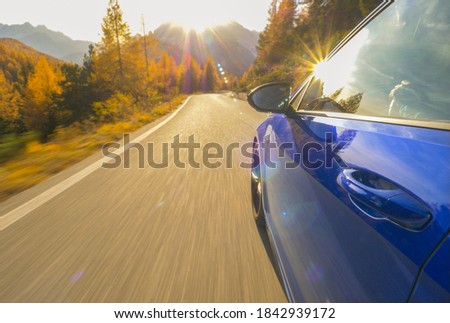 CLOSE UP, LENS FLARE: Idyllic view of sunlit Dolomites as blue sportscar drives down scenic road. Cinematic shot of golden sunbeams reflecting in the shiny door of a car cruising down mountain road.