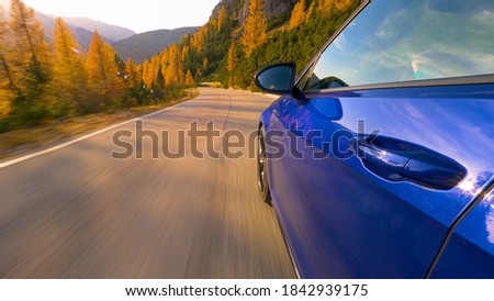 CLOSE UP, LENS FLARE: Brand new sportscar speeds down an empty mountain road in the Italian Alps at sunset. Golden fall sunbeams shine on a shiny blue car driving along scenic route in the Dolomites.